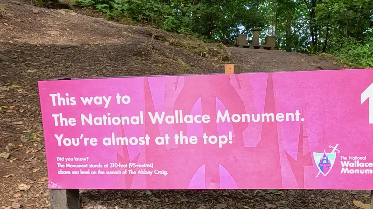A dirt trail with a sign indicating proximity to the Wallace Monument, surrounded by lush greenery.