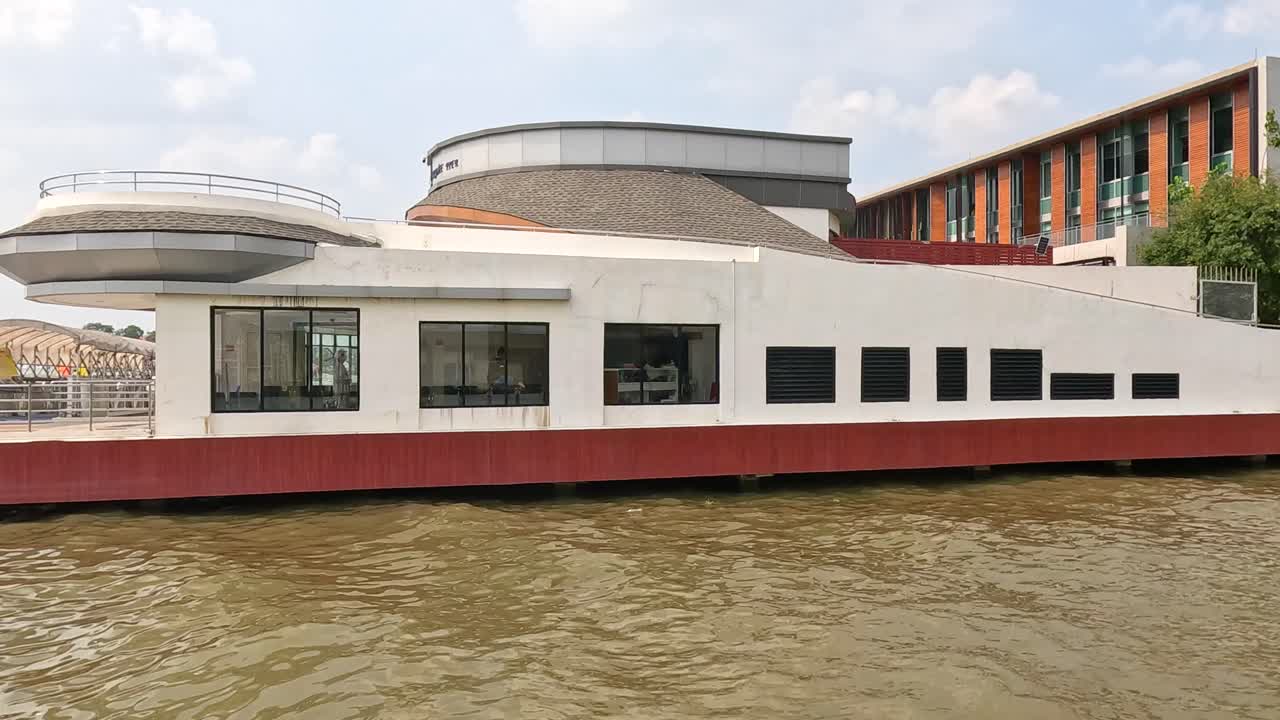 A modern building with circular architecture by a river in Bangkok, captured in bright daylight with calm water