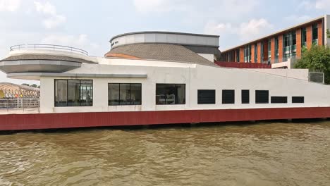 a modern building with circular architecture by a river in bangkok, captured in bright daylight with calm water