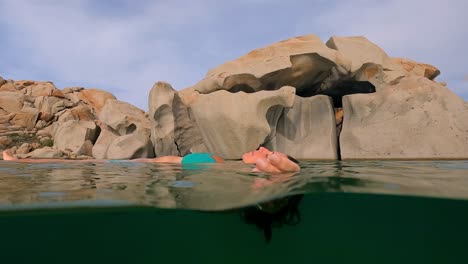 half underwater scene of woman in blue bikini relaxing floating on sea water of cala della chiesa lagoon with eroded granitic rocks in background of lavezzi island in corsica, france