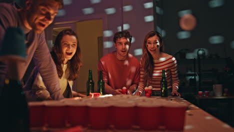 friends playing beer pong at home party with flashing lights. man throwing ball