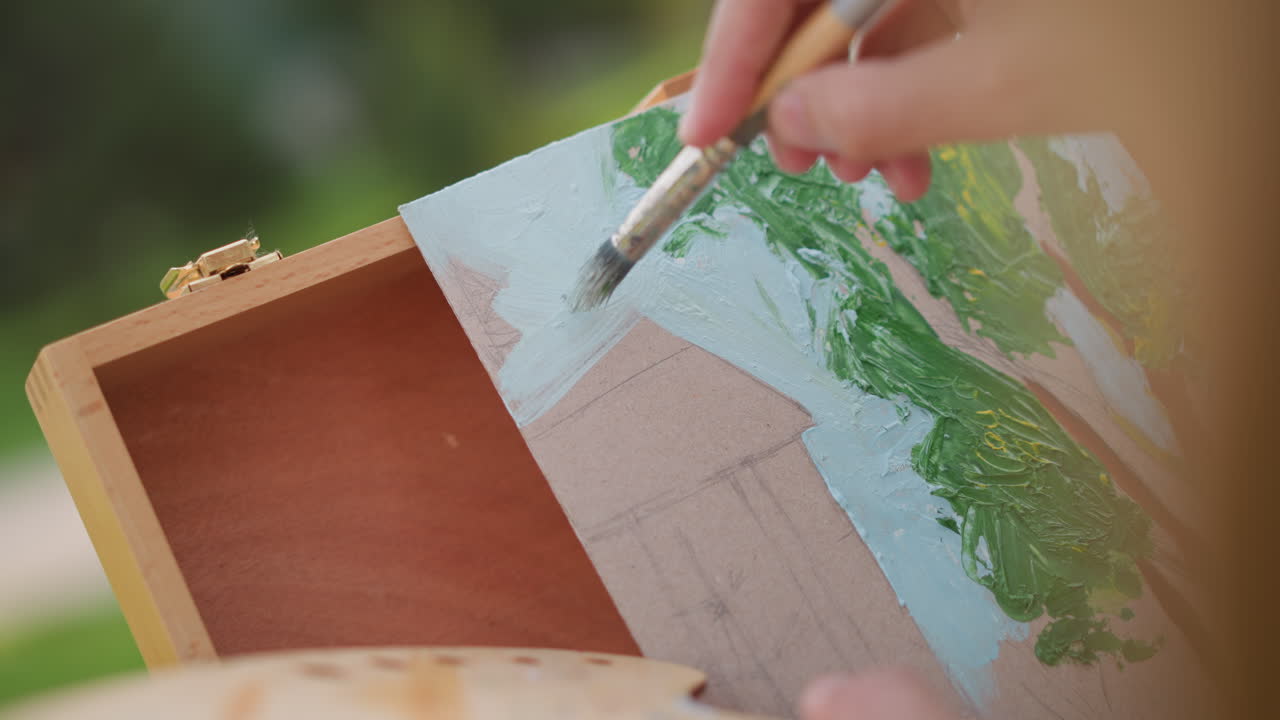 Nærbillede af kunstners hånd, der maler himlen, udendørs pleinair-session, der viser tidlig skitse af hus og træer på lærred, blide penselstrøg, tekstureret palet, varmt sollys, rolig parkstemning, hobbyist