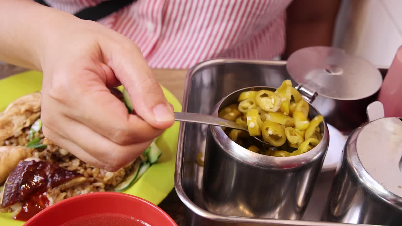 A person in a striped shirt uses a spoon to serve pickled green chili from a metal container onto a plate of Hainanese chicken rice under bright indoor lighting