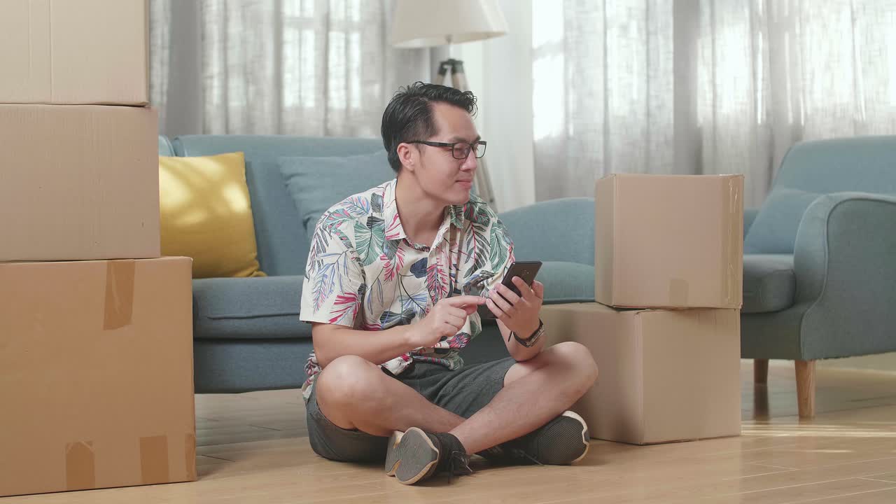 Asian Man With Cardboard Boxes Sitting On The Floor And Comparing The House To The Photo On Smartphone After Moving Into A New House