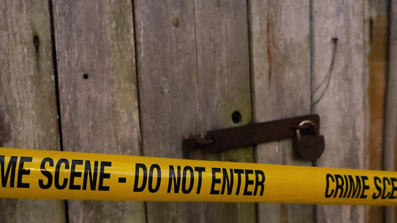 Yellow colored forensic science police crime scene tape pulled in front of an old wooden barn or countryside door with a rusty metal padlock that is locked. Video slowly gliding in front of the area