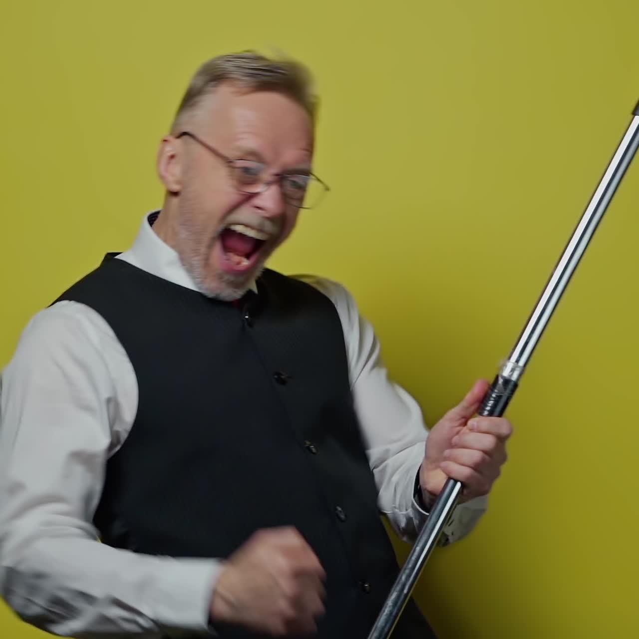 Funny elderly man dancing with a mop. Amusing old man in suit singing happily with a stick like playing the guitar on yellow background. Positive emotions.