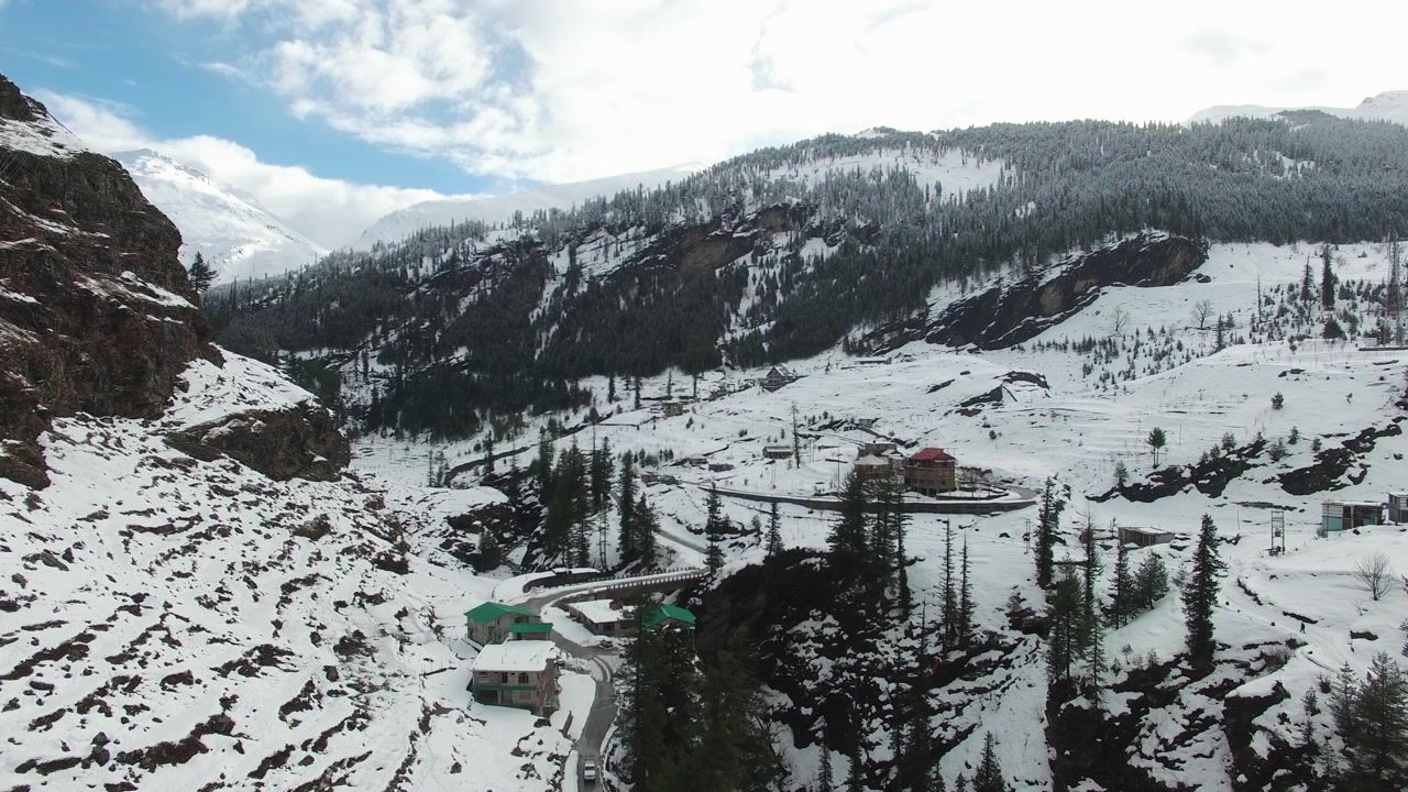 Aerial drone view over Gulaba village near Manali in Himachal Pradesh after heavy winter snowfall. Snow blankets the valley, scattered conifers, and winding mountain road under clear blue sky.