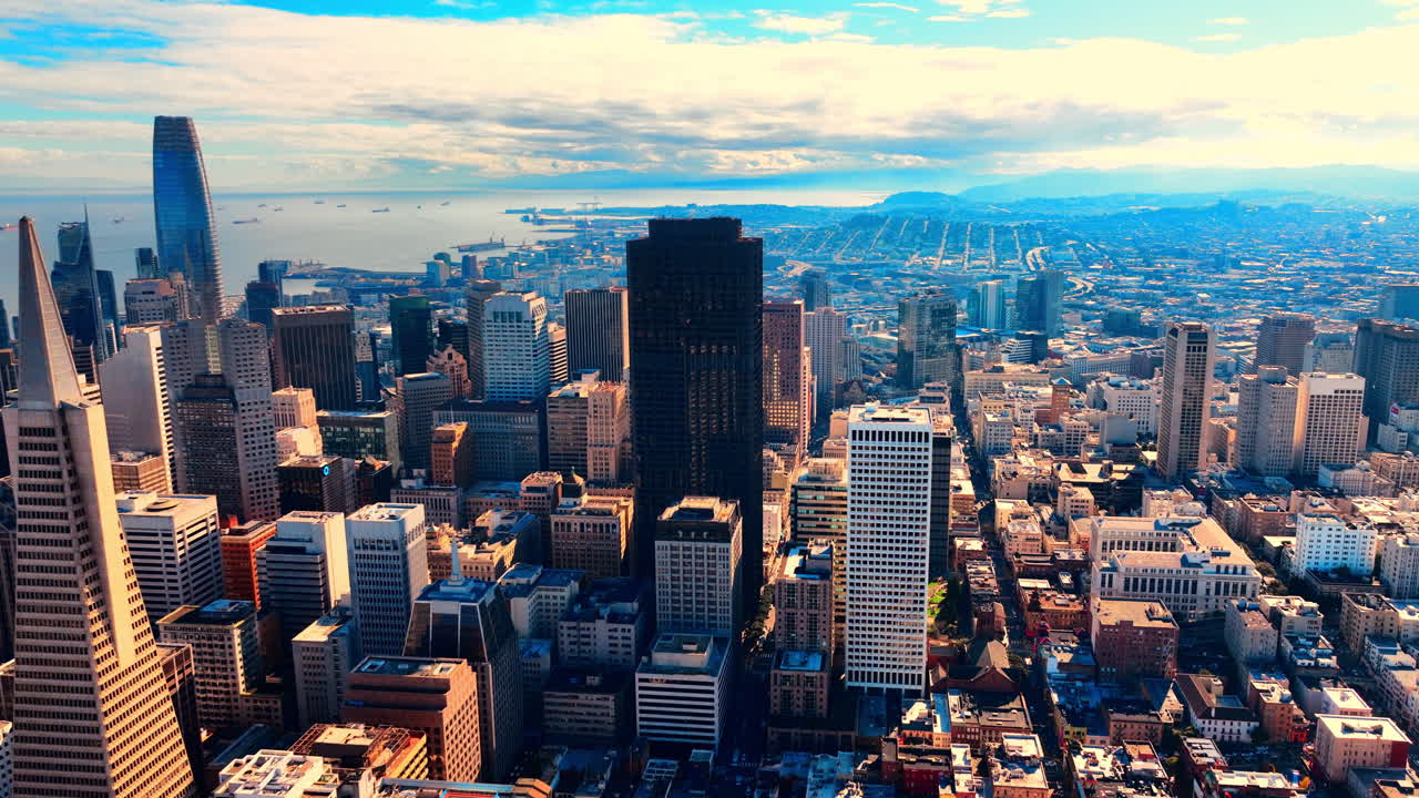Vista aérea elevada do centro financeiro de São Francisco. Arranha-céus, incluindo a Pirâmide Transamerica, cercados por edifícios residenciais e a Baía de São Francisco ao fundo, com navios.