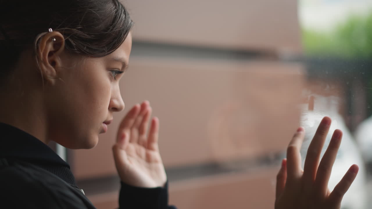 Giovane donna che preme i palmi contro il finestrino, postura da pendolare, spallina dello zaino visibile, striature di pioggia, sguardo fisso verso la strada, tranquilla pausa urbana, illuminazione cupa e nuvolosa, scatto di profilo intimo.