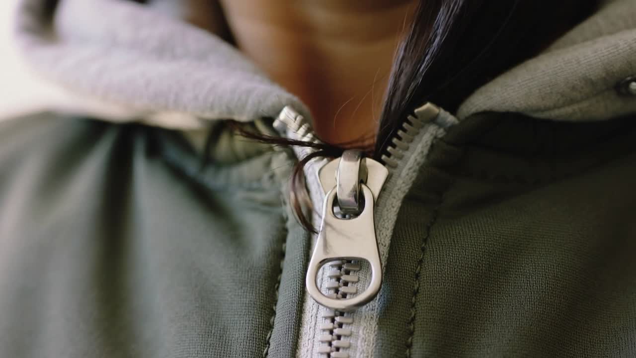 Cabelo preso no zíper de metal, mulher puxando o queixo para soltar a mecha no peito em close-up, jaqueta verde com capuz.