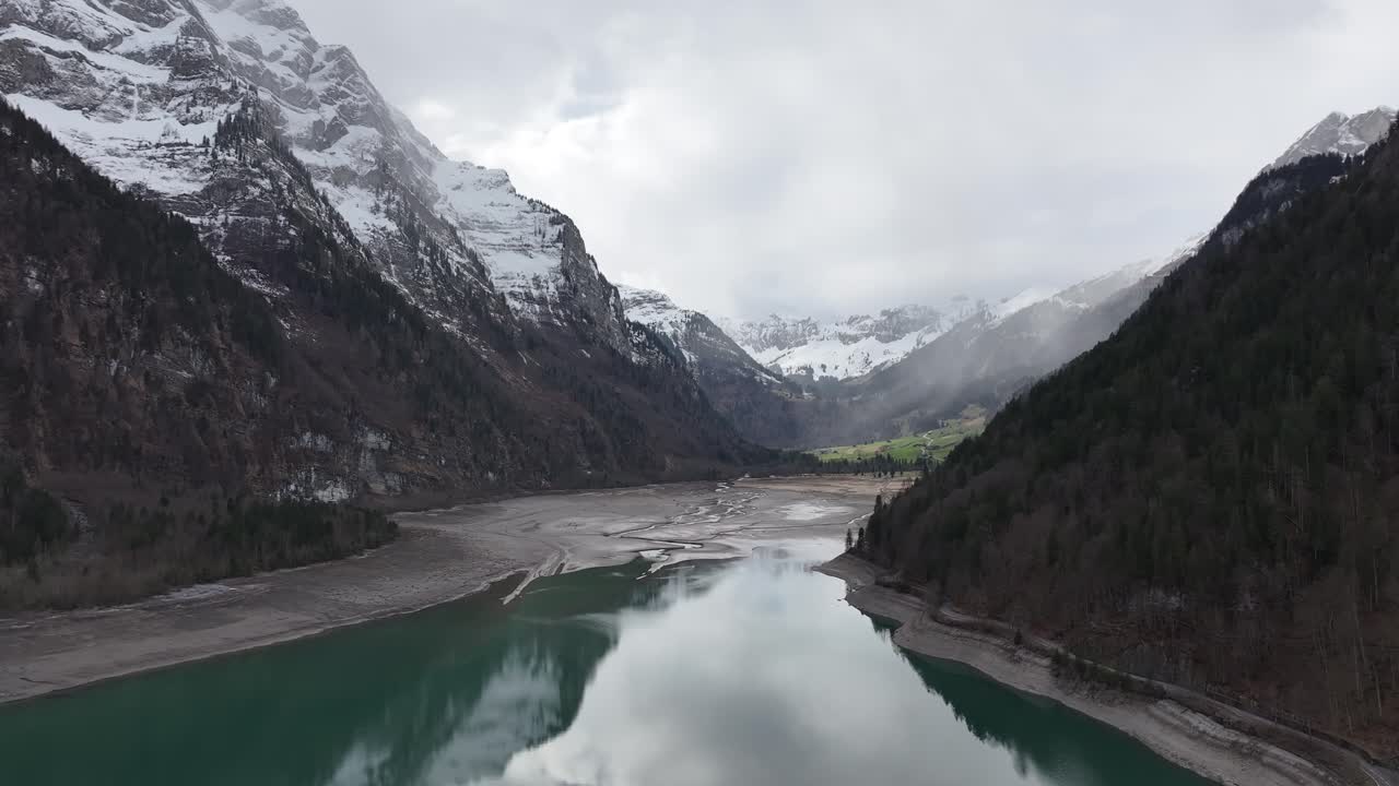 Veduta aerea del Klöntalersee con un lago turchese riflettente incastonato tra cime montuose innevate e ripidi pendii boscosi