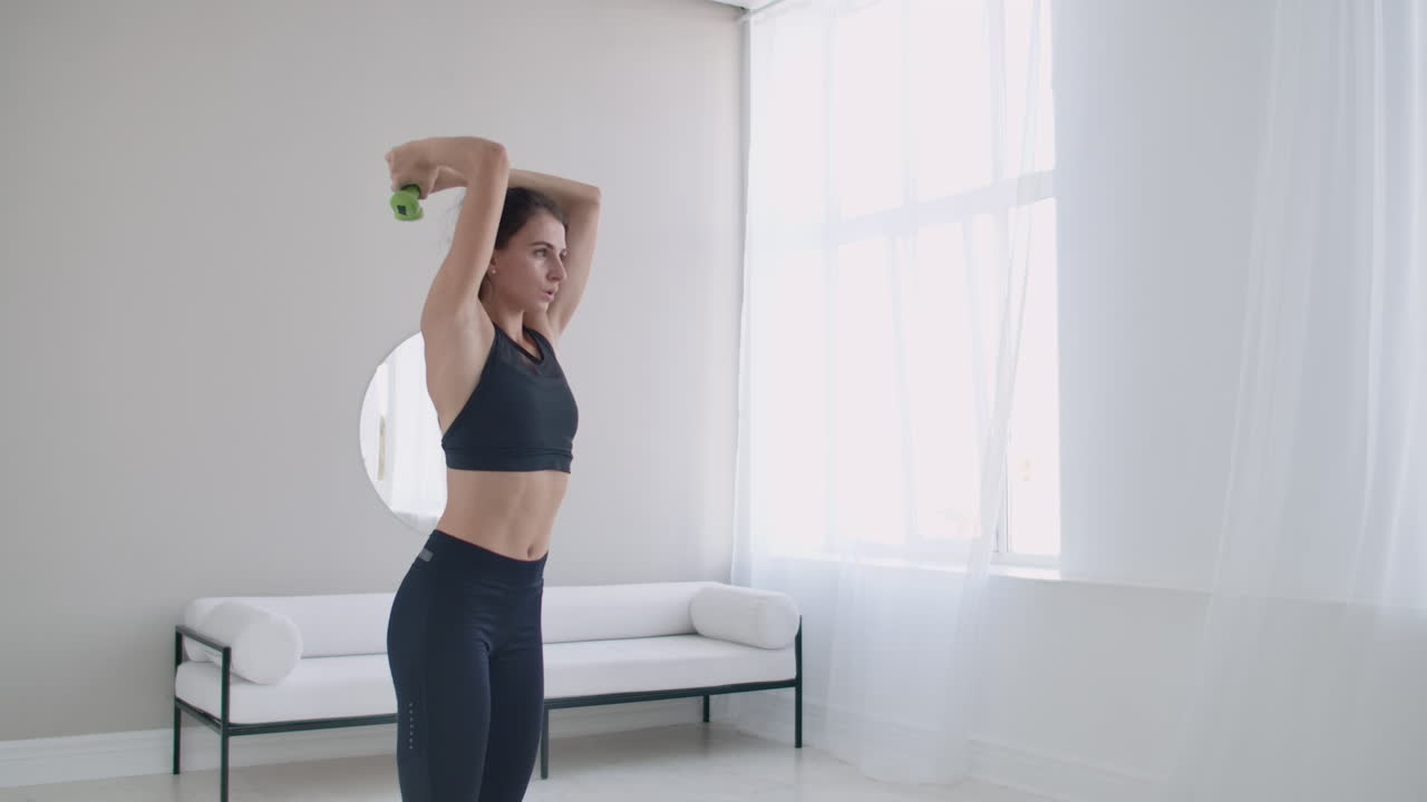 트라이세프스 근육을 위한 운동: 머리 뒤에 덤벨을 들어 올리는 운동 - 젊은 여성이 집에서 훈련하고 있습니다.