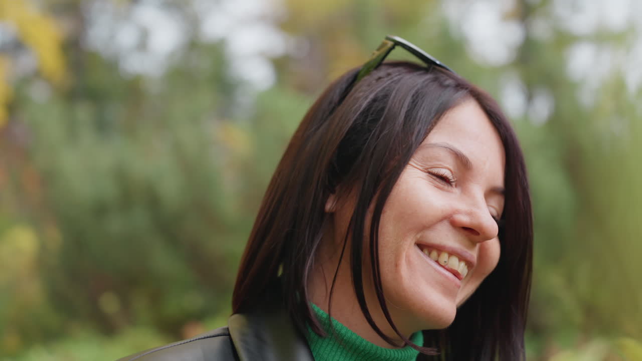 Mujer caucásica sonriente con chaqueta de cuero, gafas de sol en la cabeza y cuello alto verde, risa espontánea en un bosque con primer plano de hojas borrosas, luz cálida de otoño, retrato relajado de estilo rock