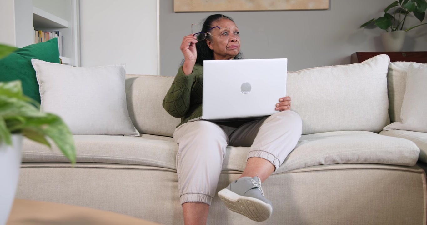 Senior African woman sitting on tan sofa, using white laptop, glancing right adjusting eyeglasses