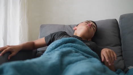 restless kid sleeping on couch covered in blanket adjusting for comfort with tissue nearby and glass cup on table. indoor relaxing environment with soft natural light streaming through window