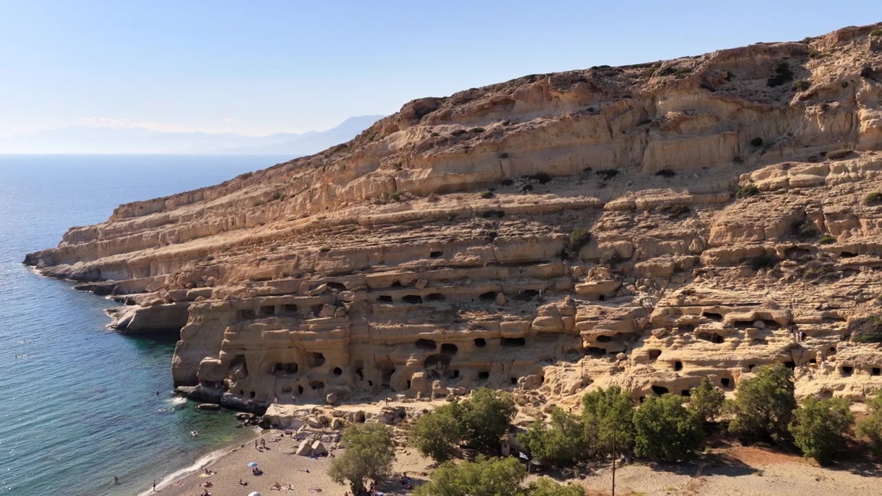 Vista aerea con drone di una vasta scogliera di arenaria costellata di antiche grotte sopra il mare limpido e turchese, Matala, Grecia