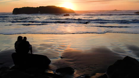 two people enjoying a beautiful sunset on the beach with a large sea stack in the distance which the sun is setting behind