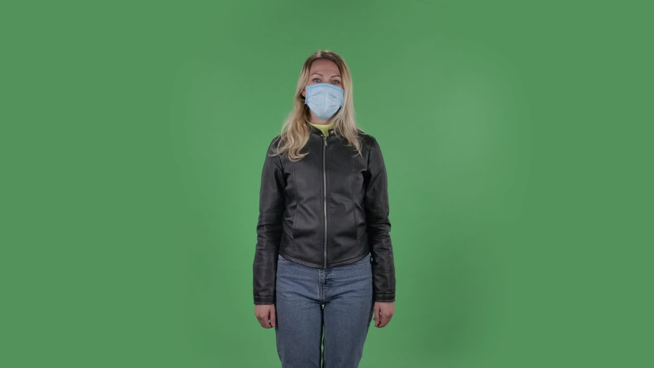 Portrait of beautiful young woman in medical mask is looking at camera and joyfully making sign ok. Blonde with loose hair in a black jacket and jeans on a green screen in the studio. Health Protection Corona Virus Concept