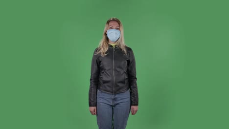 portrait of beautiful young woman in medical mask is looking at camera and joyfully making sign ok. blonde with loose hair in a black jacket and jeans on a green screen in the studio. health protection corona virus concept