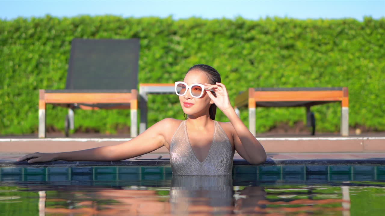 Smiling Asian woman with white swimsuit and vintage sunglasses relaxing in pool water