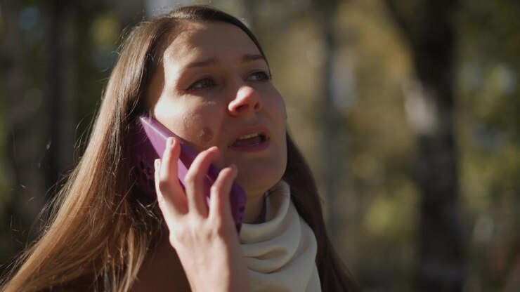 Close up of woman holding and using purple phone outdoors with sunlight filtering through trees. Her long brown hair is visible wearing light colored scarf around her neck. background shows