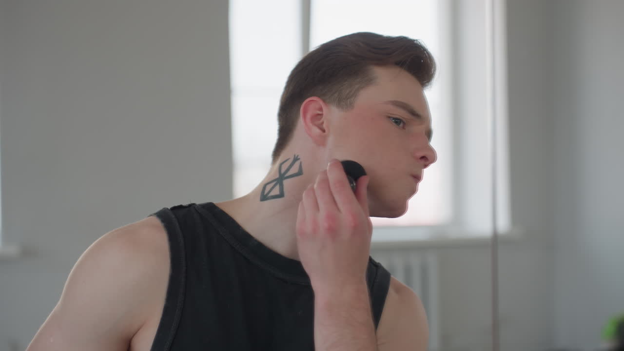 mirror shows neck tattooed boy gently applying makeup with sponge under soft natural light from window in bright minimalist studio reflecting urban building beyond over cushioned stool