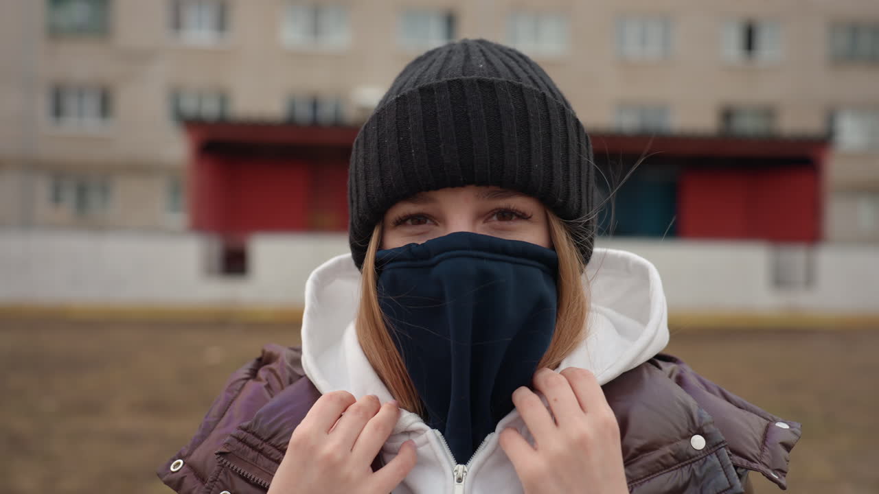 후드와 따뜻한 장비를 착용한 여성, 편안함을 보여주는 동네 배경에서 얼굴을 가린 여성, 후드 재킷과 마스크를 착용하고 안전하고 평온한 야외 환경을 즐기는 여성