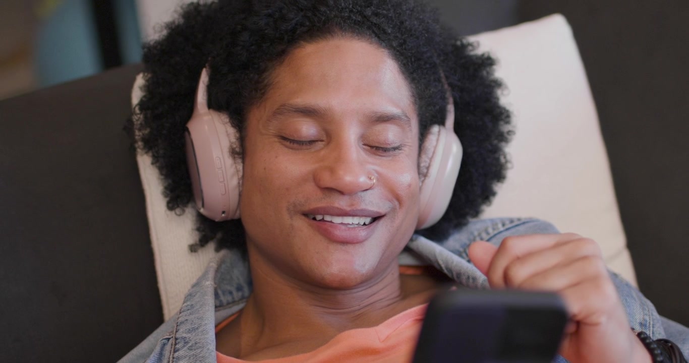 Uomo afroamericano sdraiato a casa con cuffie rosa che sente un avviso, tocca il telefono e legge messaggi