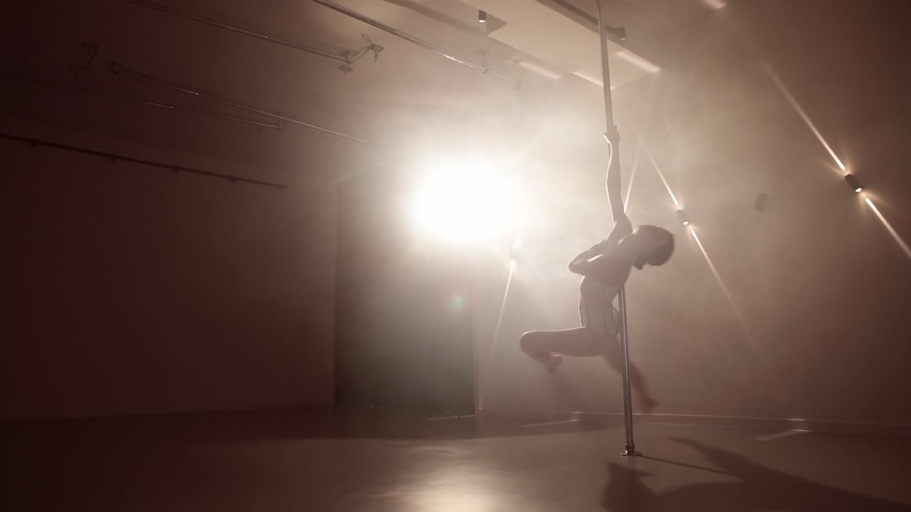 Young female artist practicing pole dancing in a smoke-filled room