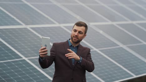 Porträt-Eines-Gutaussehenden-Mannes-In-Einer-Jacke-Mit-Einem-Tablet,-Der-Bei-Einem-Videoanruf-Auf-Dem-Hintergrund-Eines-Solarkraftwerks-Spricht
