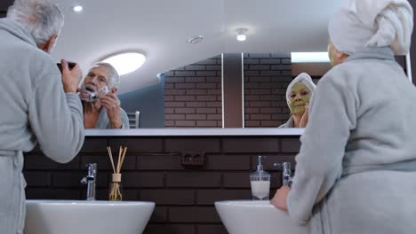 Old-senior-couple-man-and-woman-doing-morning-hygiene-procedures-and-looking-into-a-mirror