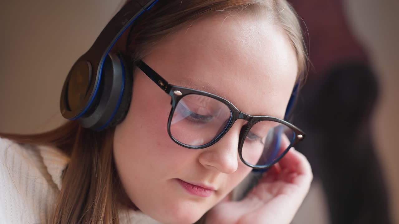 close up of focused woman wearing headphones and glasses gently moving head while listening to music with soft ambient lighting and blurred wall art in background