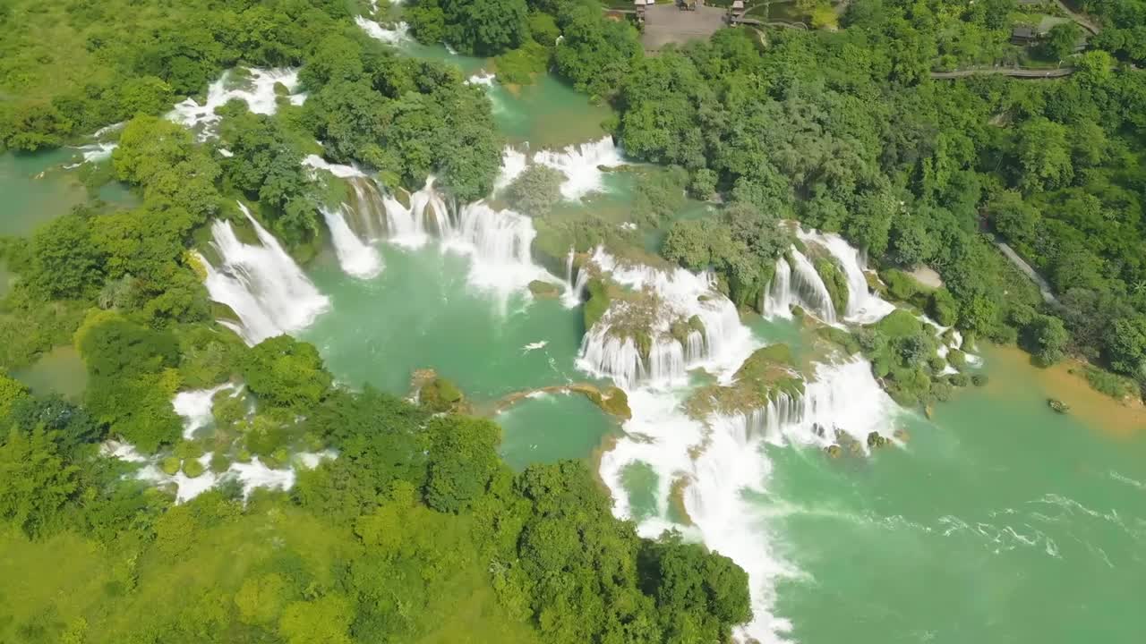 A wide aerial view of a multi-tiered waterfall surrounded by lush green mountains, a turquoise river, and a sandy beach with small boats and visitors below.