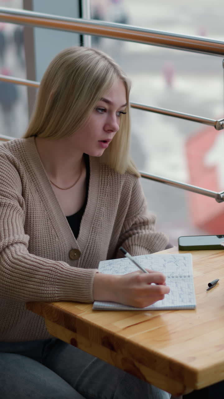 Student thoughtfully writing with pen and paper while enjoying coffee, deep in concentration, with urban winter background visible through window in cozy cafe setting