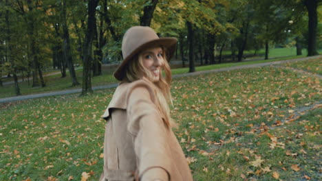 rear view of caucasian young blonde woman in a hat and coat walking in the park and leading the camera with her, then she turns and smiles