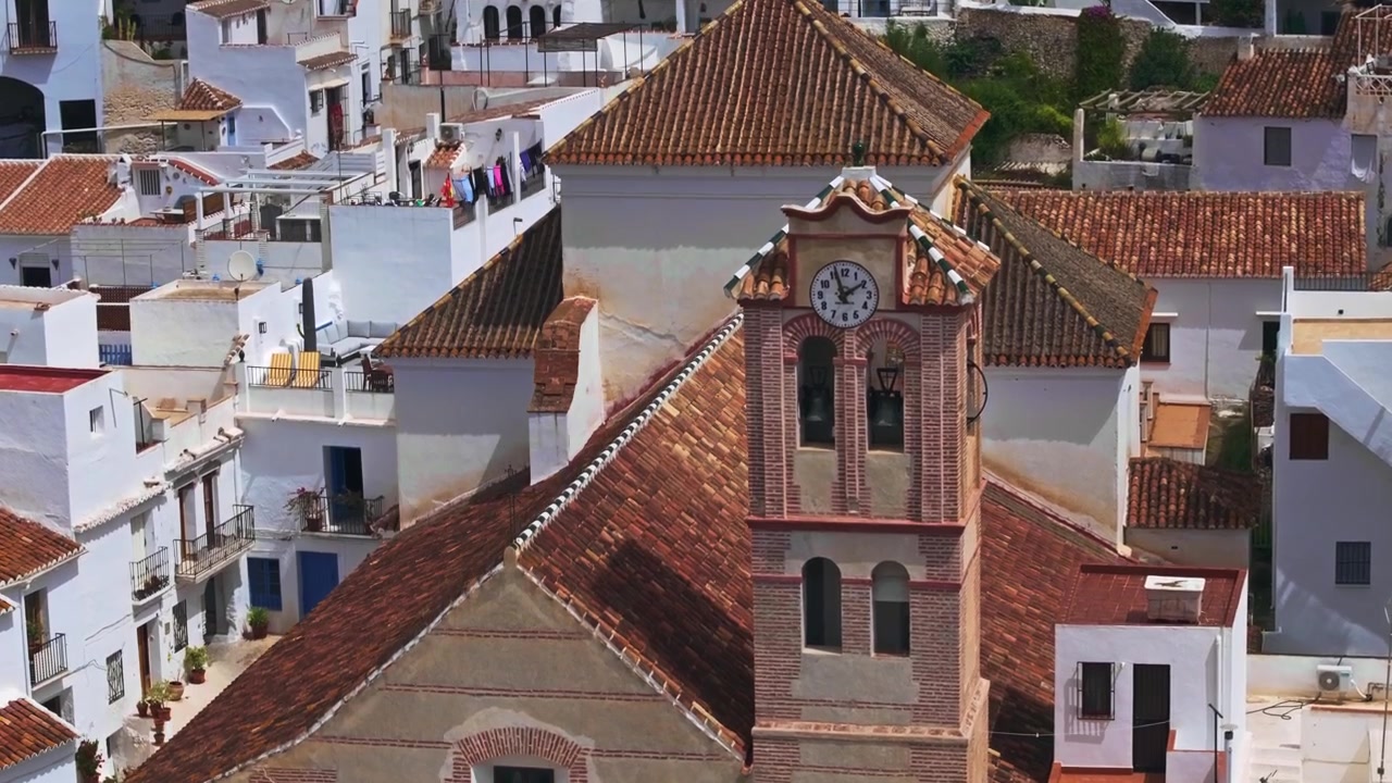 Frigiliana, Endülüs, İspanya'dan, badanalı yamaç evleri ve dağ manzarasıyla çevrili bir kilise kulesinin yükselen havadan görünümünü gösteren drone görüntüleri.