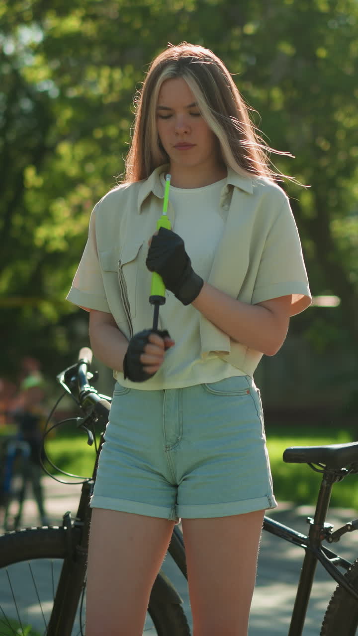 joven con cabello rubio se encuentra a la luz del sol, enfocado mientras sostiene la bomba de aire verde al lado de la bicicleta, el fondo muestra una figura borrosa caminando y árboles exuberantes