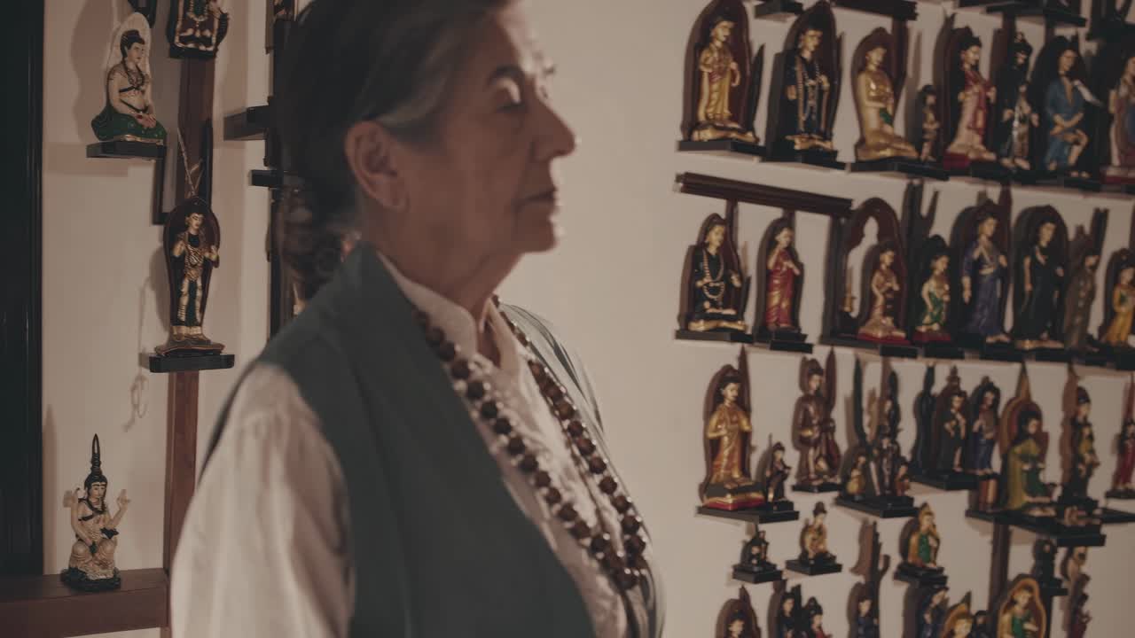 Elderly woman wearing a vest and a necklace is inside a house, looking at a wall full of religious figurines, with a wooden cross visible on the left and a framed picture on the right