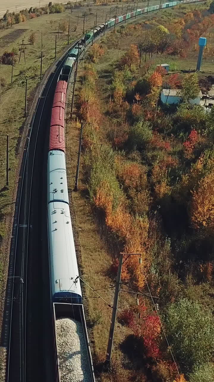 a long freight train with building materials is moving by railway on the background of fields and forests in the summer on a sunny day. Aerial view Vertical video