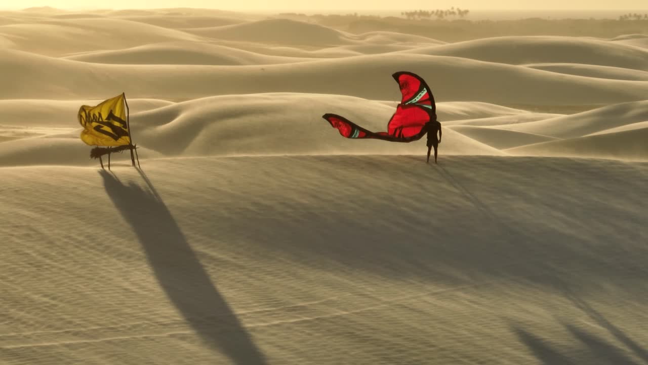 바람이 부는 모래와 밝은 황금빛 햇빛이 있는 모래 언덕 꼭대기에 붉은 초승달 연을 들고 노란색 깃발 옆에 서있는 사람의 매우 극적인 공중 궤도 샷