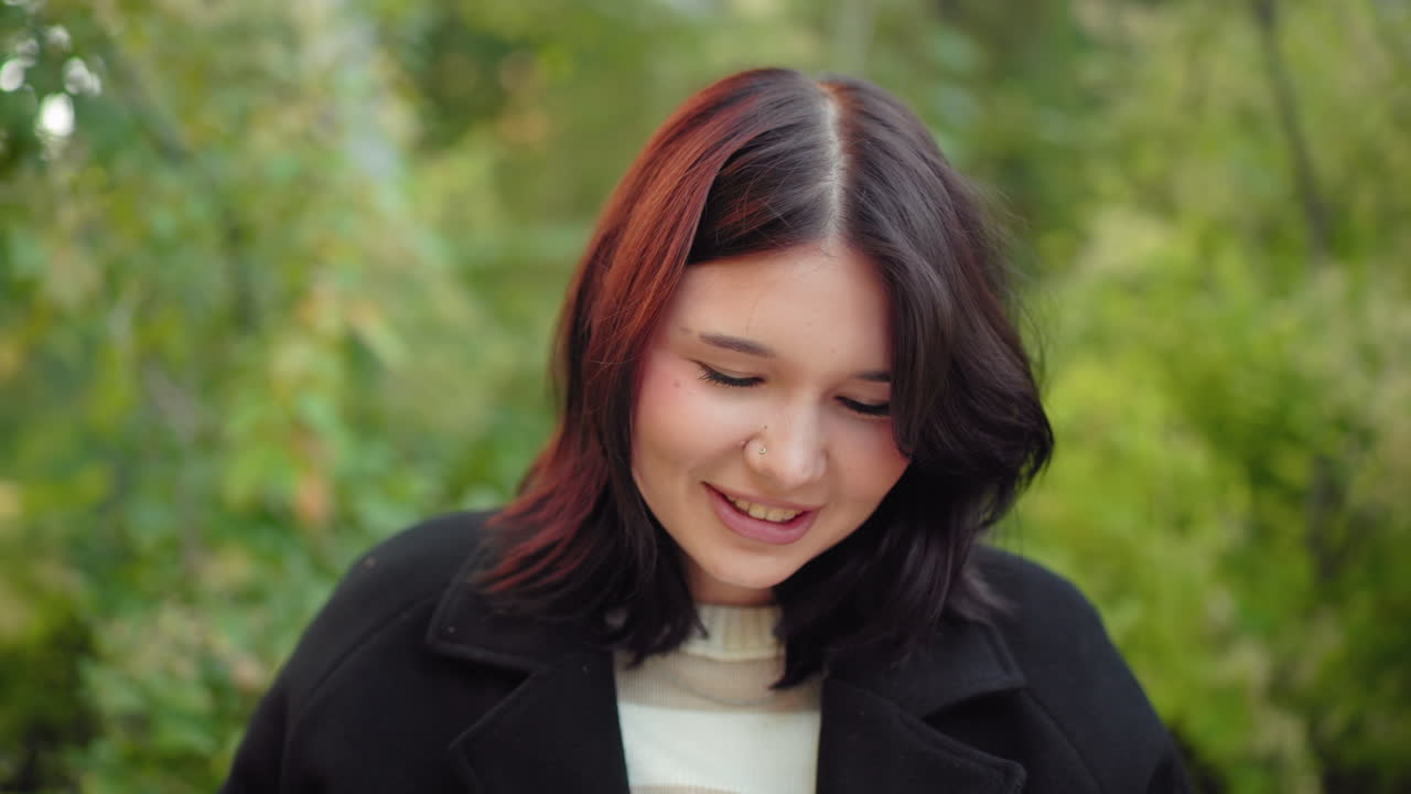Close view of nature lover smiling with eyes on book, seated in autumn arboretum, black coat and striped sweater visible, soft greenery behind, gentle light across face during outdoor reading moment
