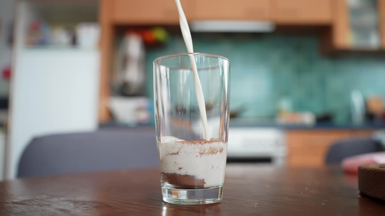 Il latte si riversa in un bicchiere trasparente sopra il cacao in polvere, creando strati cremosi e sedimenti di cacao su un tavolo di legno, con una cucina domestica leggermente sfocata sullo sfondo.