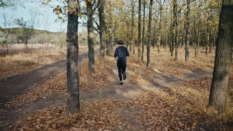 a brunette man with curly hair in a black sports uniform runs through the autumn forest along an earthen path and looks at his watch in the morning