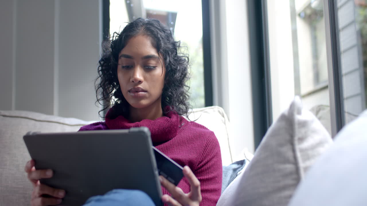 Biracial woman sitting on sofa using tablet for online shopping at home, slow motion
