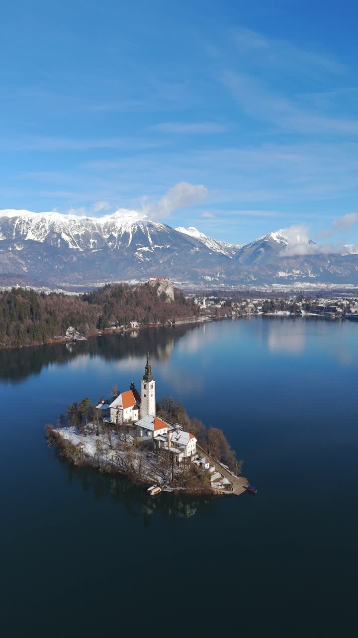 Plano de travelling lento que avanza sobre el sereno lago de Bled en invierno, revelando la emblemática iglesia de la isla con el castillo en la parte superior, rodeado de Alpes y bosques cubiertos de nieve, Eslovenia