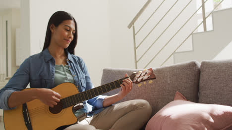 video of happy biracial woman sitting on sofa and playing guitar