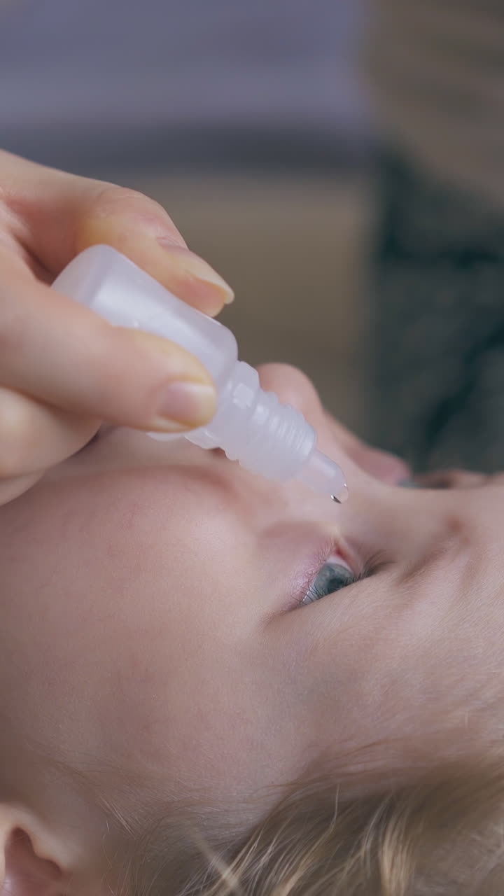 mommy gives eye drops to crying little girl lying on soft beige bed in light room extreme close view