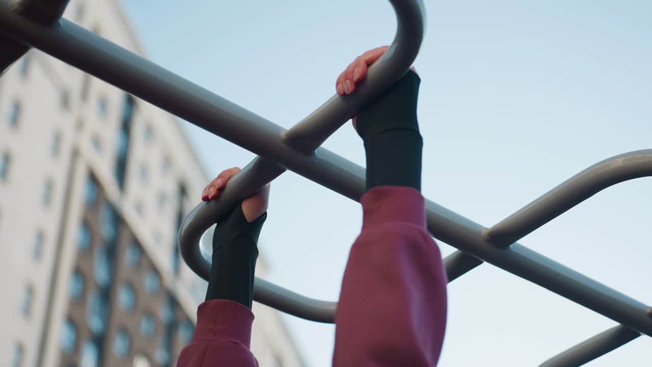 Outdoor trainer wearing black gloves grips silver monkey bars overhead against clear sky in urban park fitness area, showcasing controlled movement, strength, focus, endurance and resilience