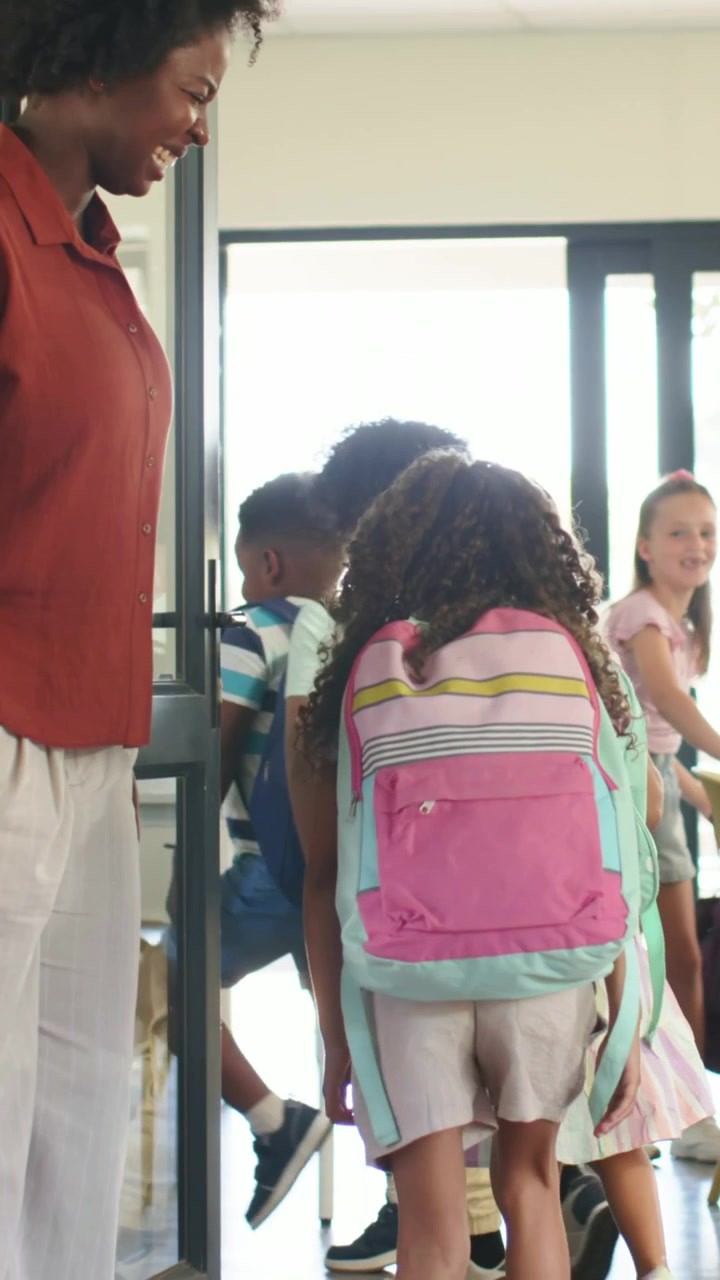 Vertical video: Teacher in rust top opening glass door, students entering with pink packs to class