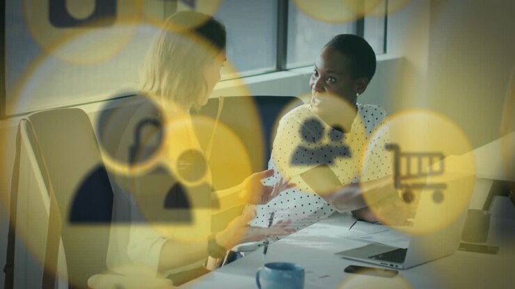 Animation of multiple icons over diverse female coworkers discussing reports on laptop in office